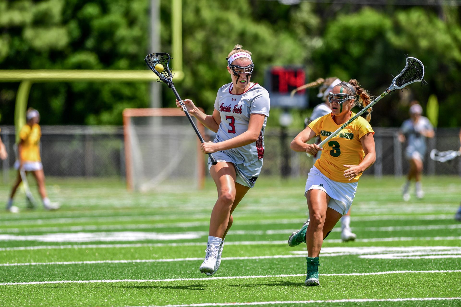 A Florida Tech women's lacrosse player in a gray uniform evading a defender from Saint Leo in a yellow uniform during a game on a green field